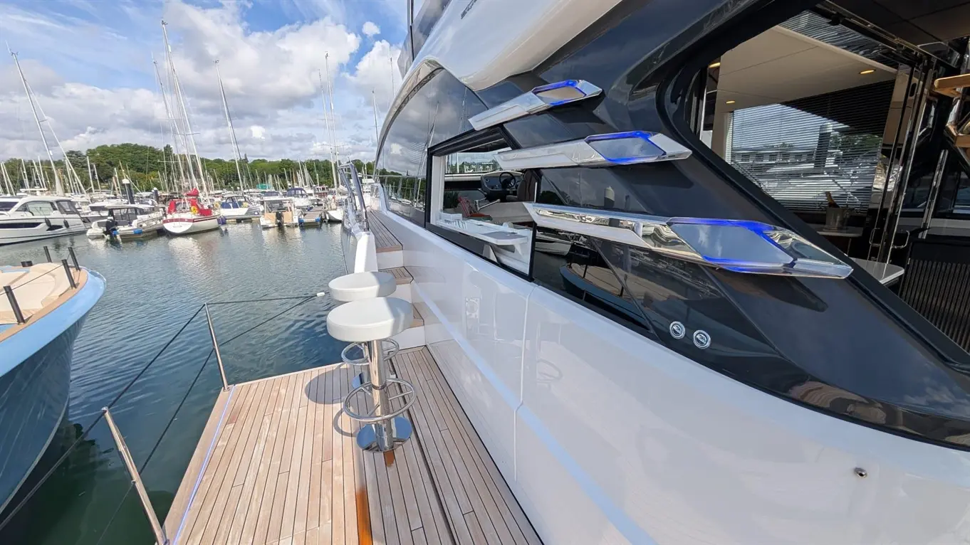 Luxury yacht with bar stools docked at a marina, surrounded by boats and reflections on water under a cloudy sky.