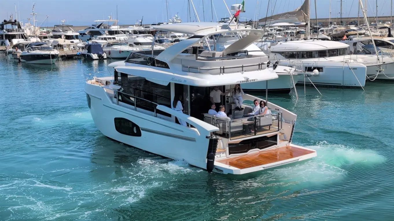 Luxury yacht with passengers enjoying the sun, surrounded by boats in a marina under clear blue skies.