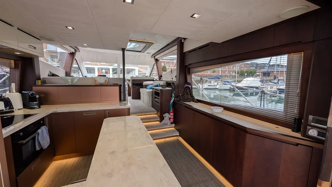 Modern yacht interior showcasing a fitted kitchen, seating area, and views of moored boats through large windows.