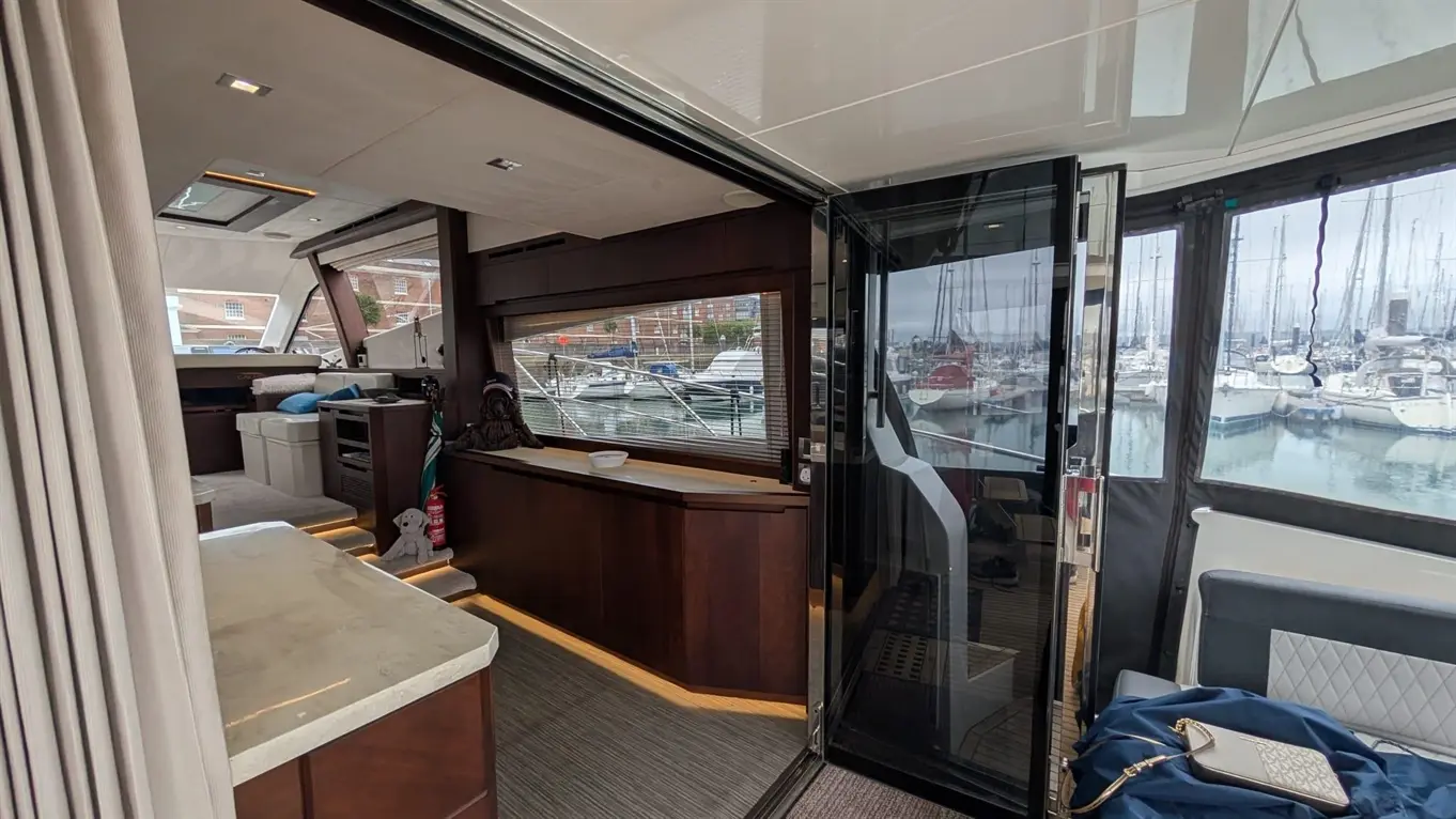 Interior of a modern yacht showing a spacious saloon with a view of the marina and boats through large windows.