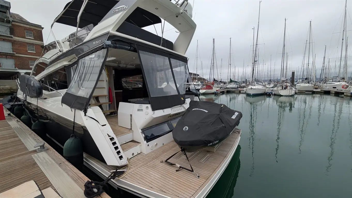 A sleek Galeon 550 yacht docked at a marina, featuring a covered jet ski and moored boats in the background on a cloudy day.