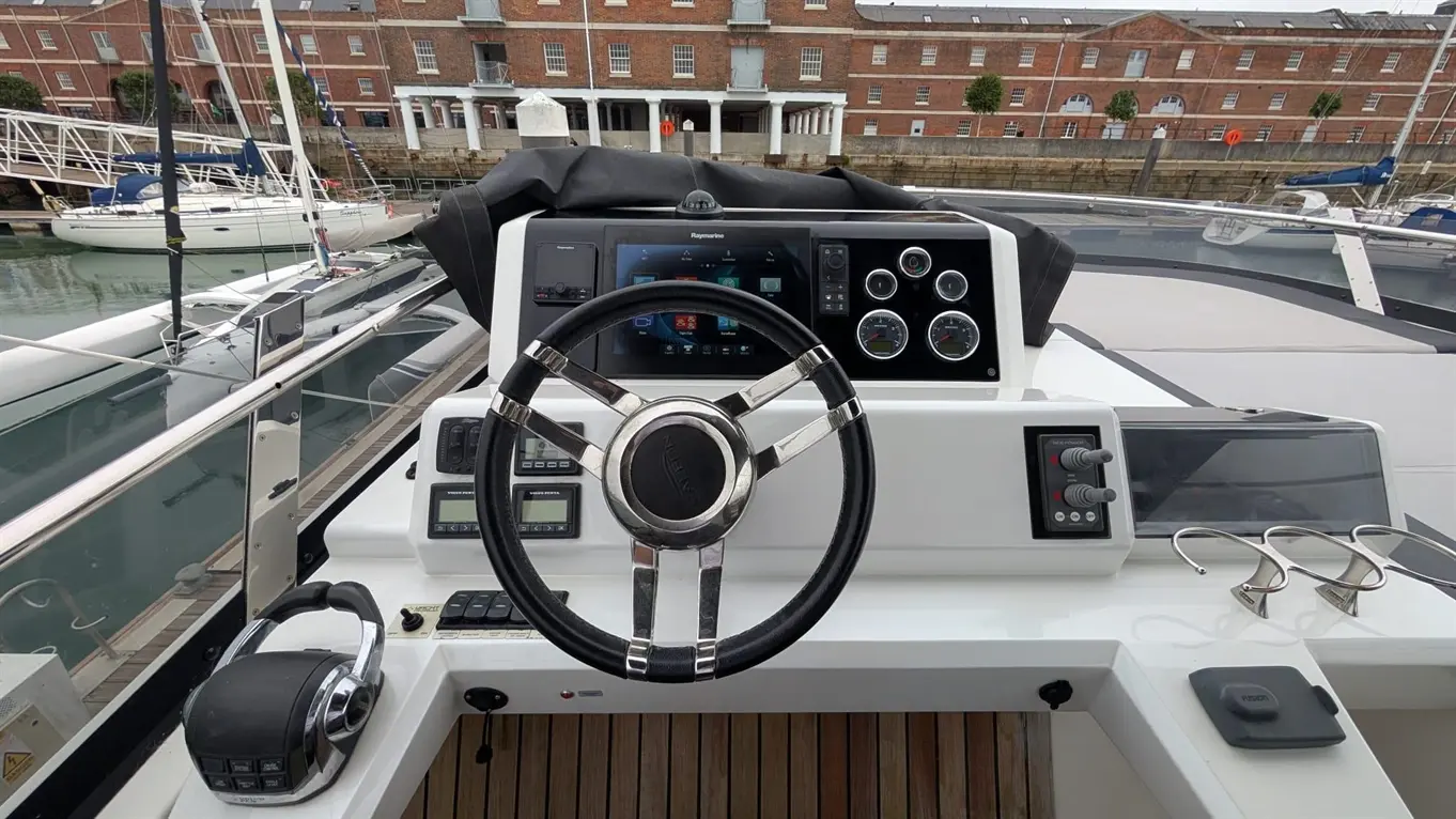 Boat cockpit view featuring a steering wheel, control panel, touchscreen display, and nautical equipment, set in a marina.