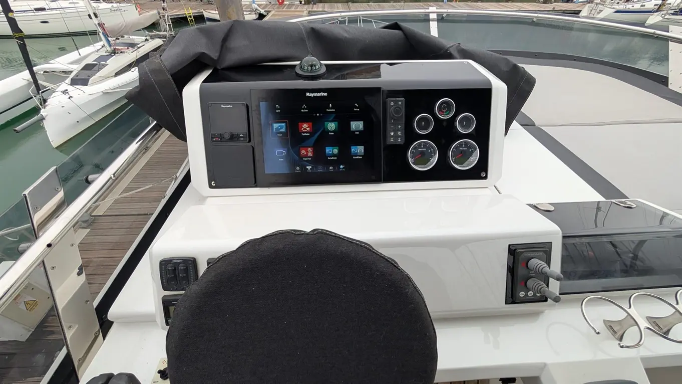 Boat cockpit with a Raymarine navigation screen, gauges, and controls. A black seat cushion is visible, with boats in the background.