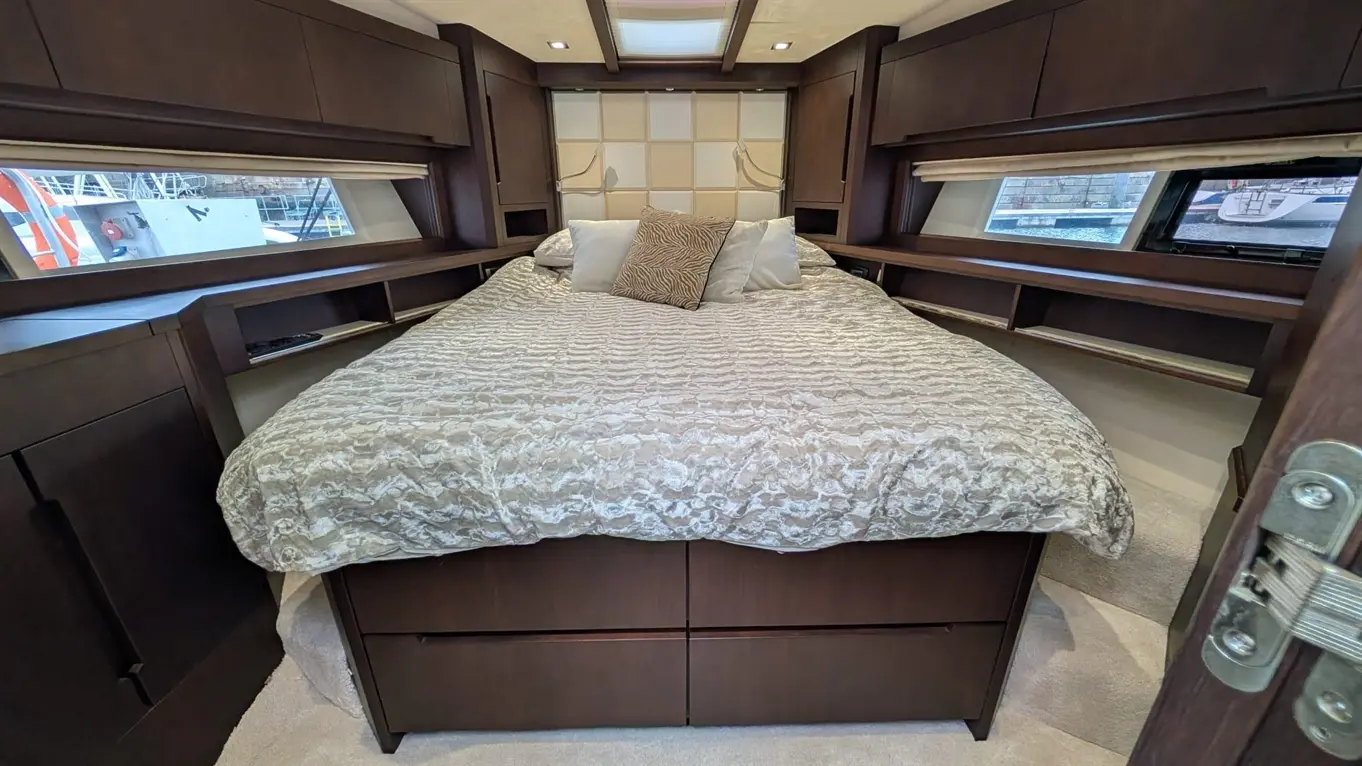 Modern boat bedroom featuring a double bed with textured bedding, storage cabinets, and a view of the marina through windows.