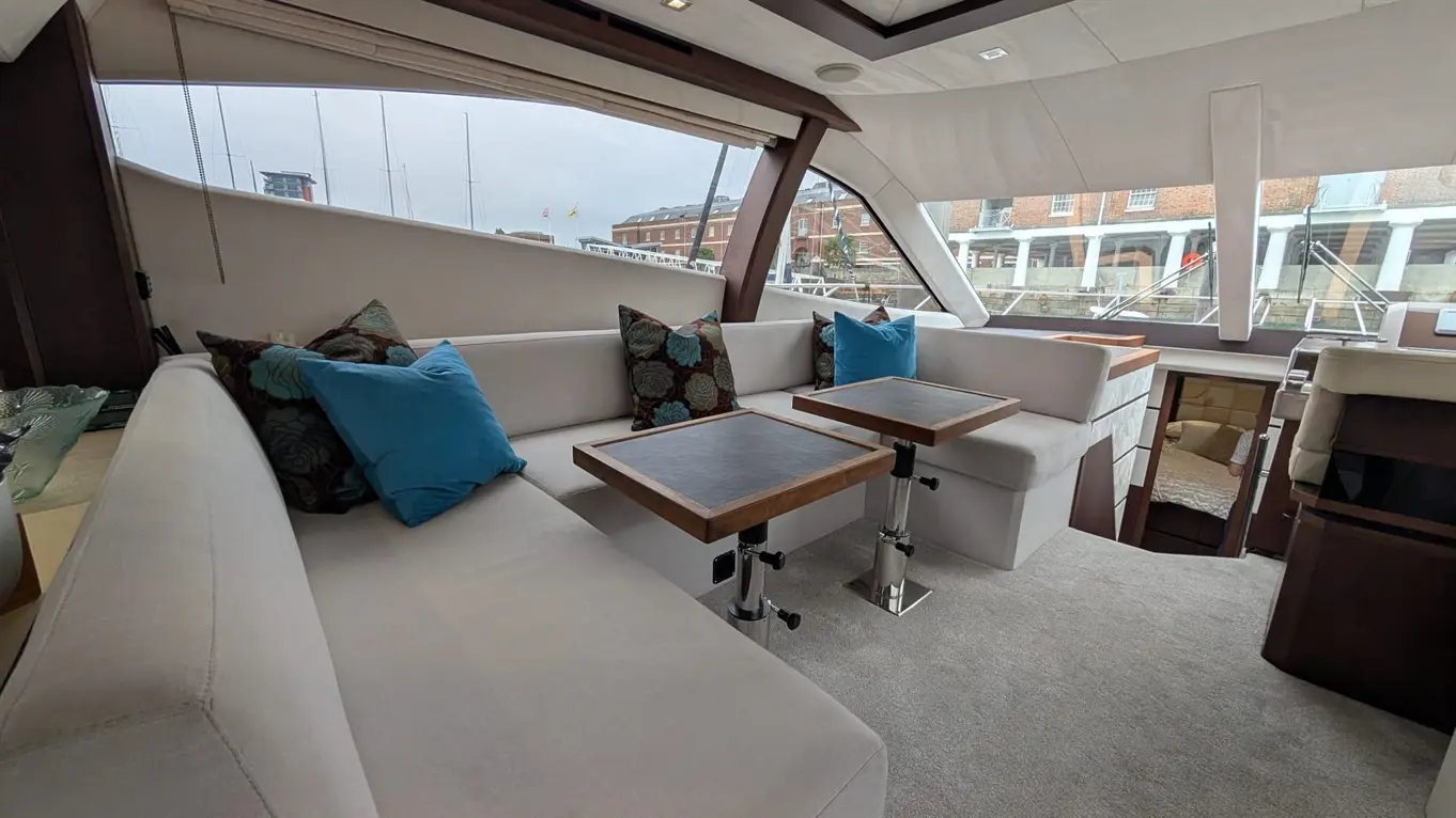 Stylish yacht interior featuring cream sofas, blue cushions, and two adjustable wooden tables, with views of the marina outside.