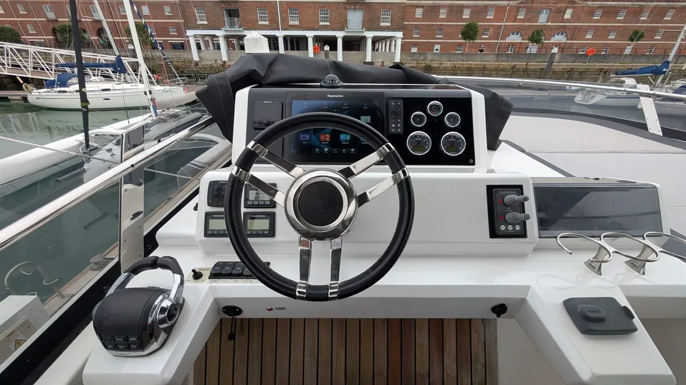 Boat interior with a steering wheel, control panel, Raymarine screen, and cloth cover, overlooking a marina with yachts.