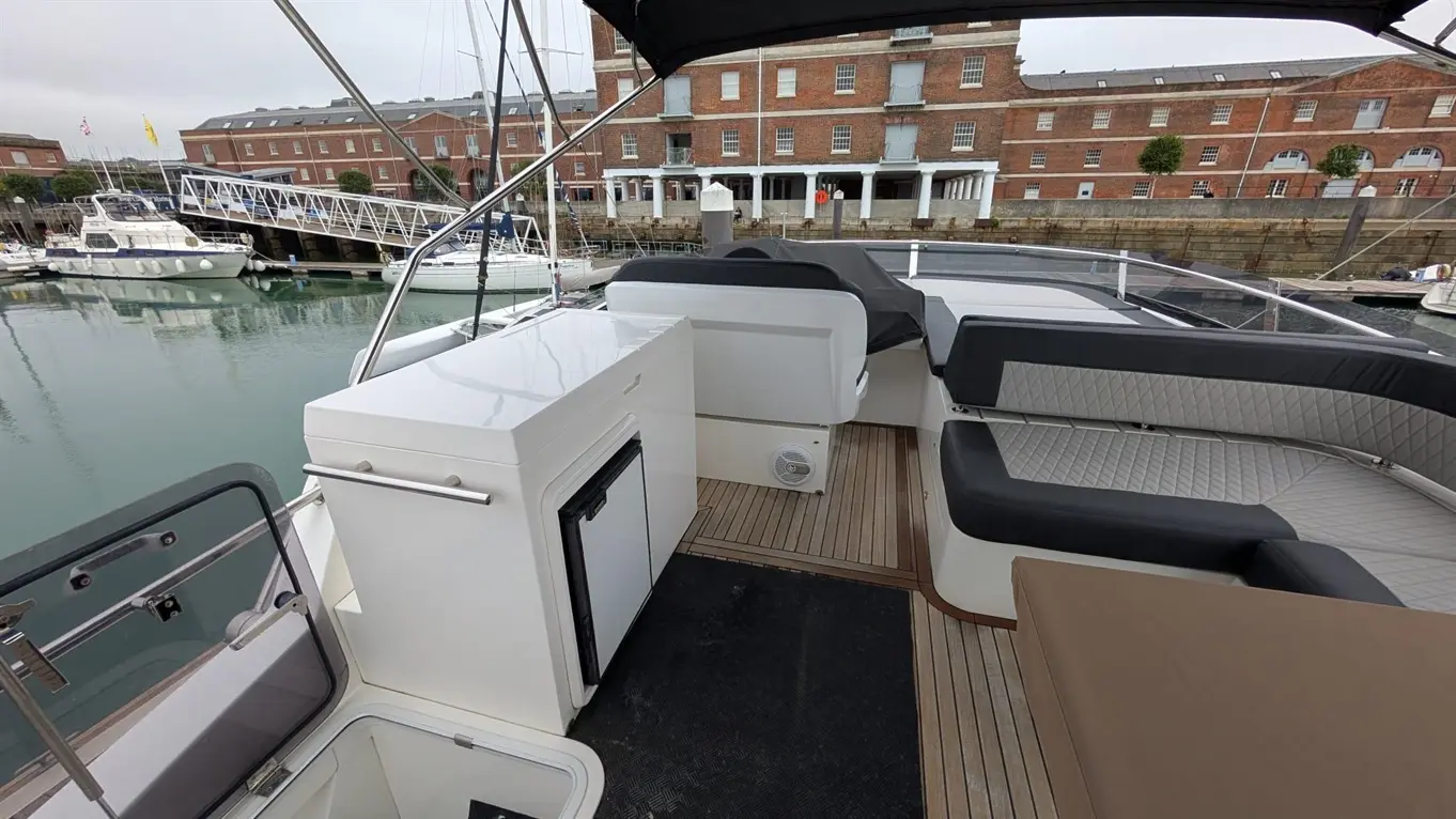 Modern boat deck with a small kitchen unit, seating area, and docked boats in the marina visible in the background.