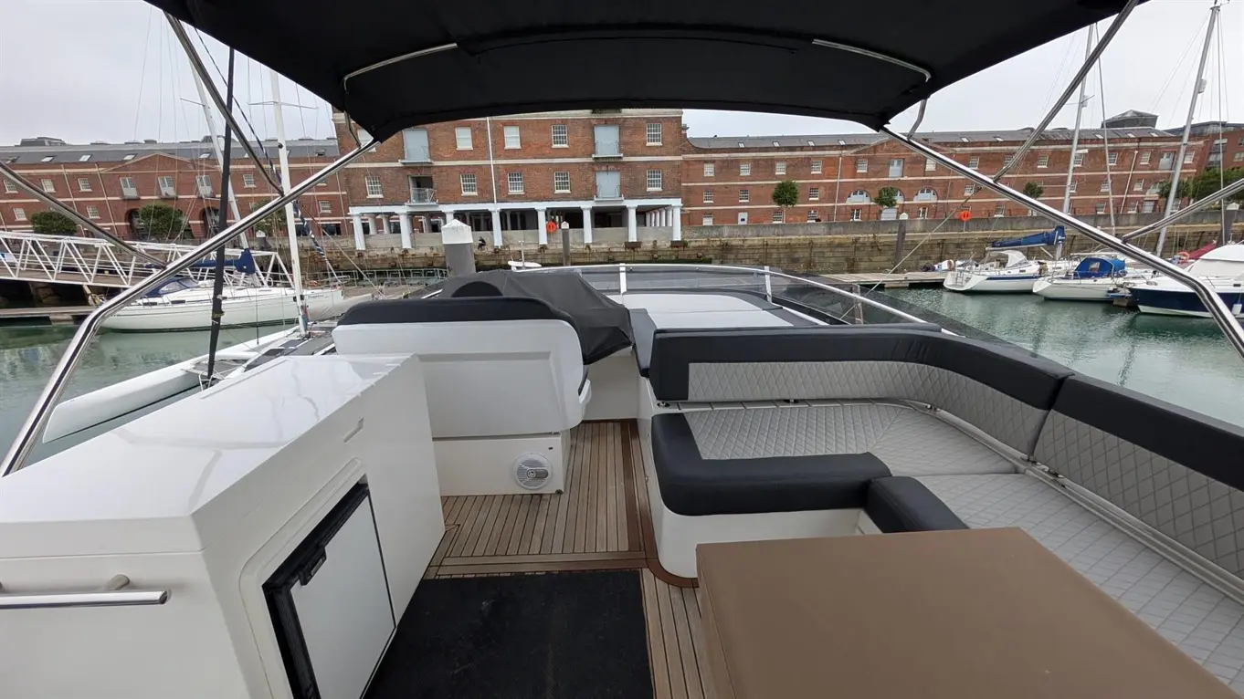 A modern boat deck featuring comfortable seating, a sunshade, and views of a marina with moored yachts and brick buildings.