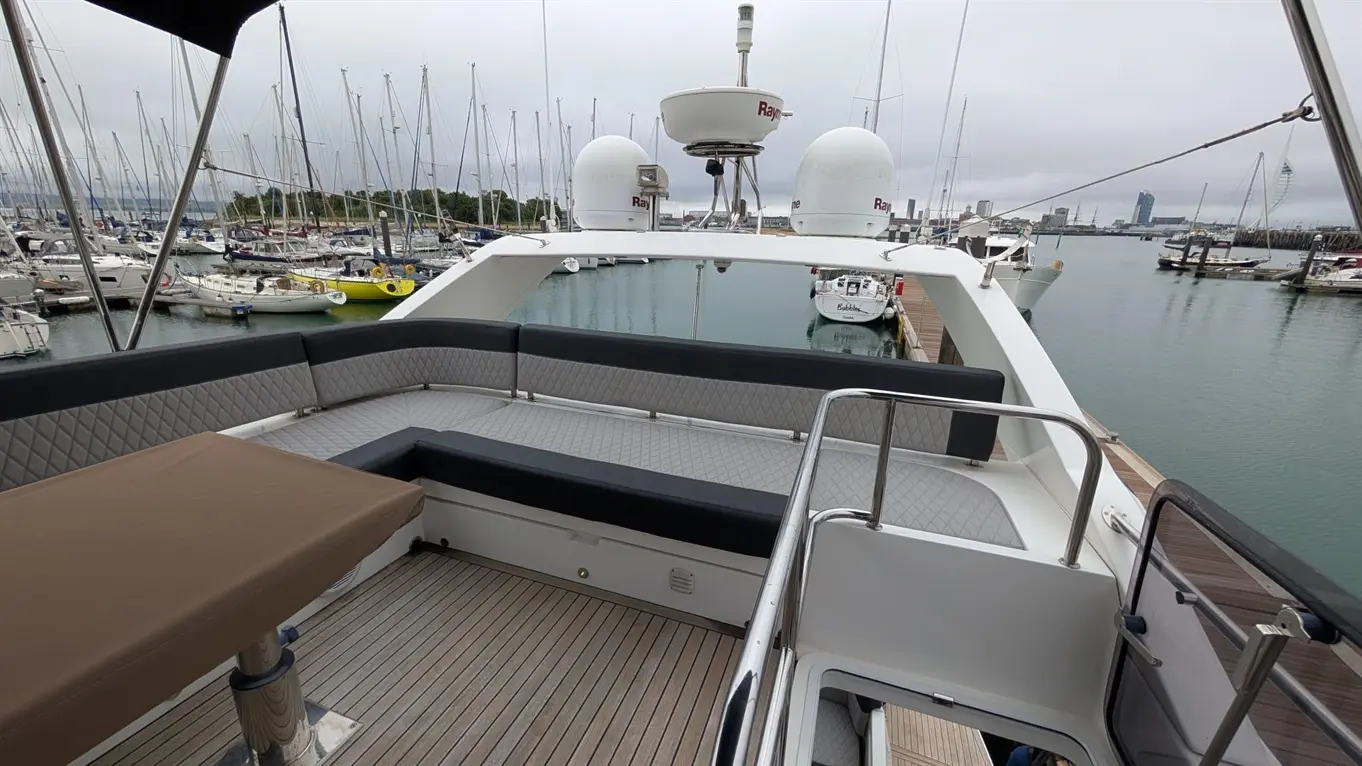 A yacht's deck features grey seating and a brown table, overlooking a marina filled with moored sailboats on a cloudy day.