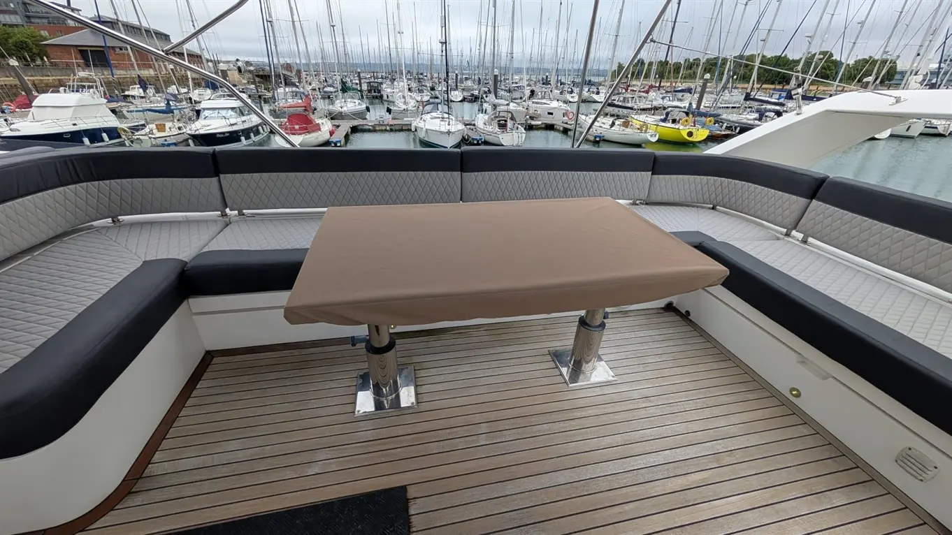 Seating area on a boat, featuring a brown table cover, grey and black cushions, with a marina filled with boats in the background.