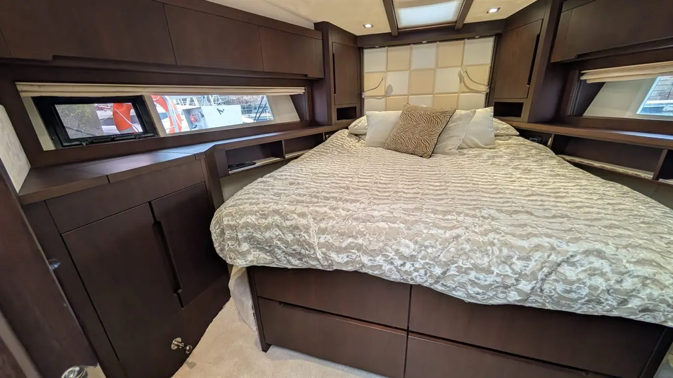Interior of a boat bedroom featuring a plush bed with decorative pillows, wooden cabinets, and a view of boats outside.