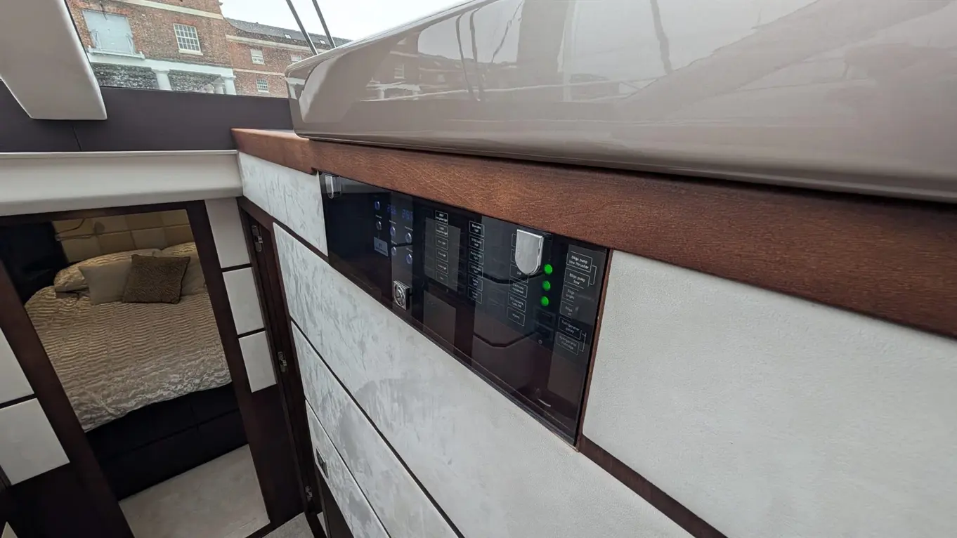 Modern boat interior featuring a control panel with illuminated buttons, leading to a bedroom with a textured bedspread.