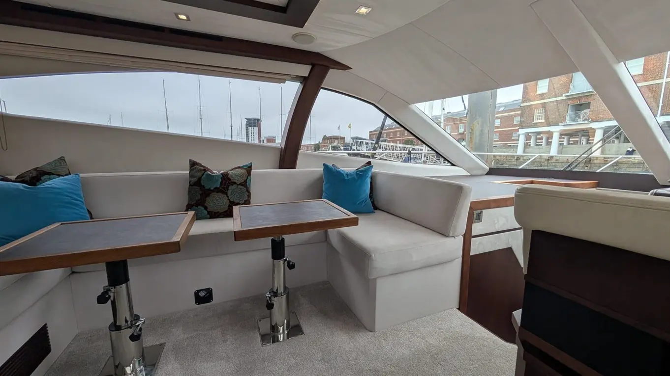 Luxurious yacht interior with plush seating, tables, and views of moored boats and historic buildings through large windows.