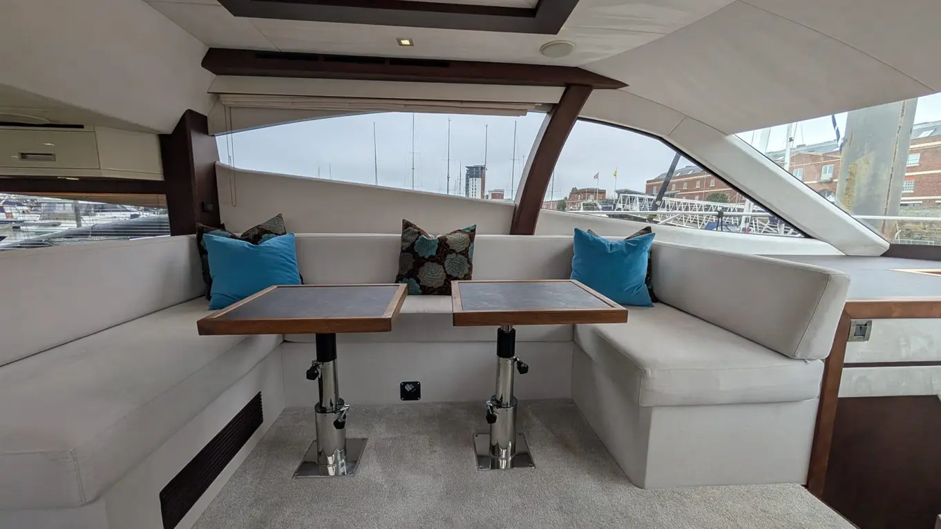 Stylish interior of a yacht featuring a sofa, two wooden tables, colourful cushions, and a view of the marina through large windows.