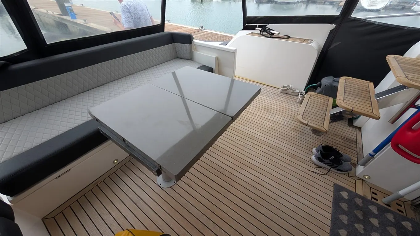 Interior of a boat featuring a grey table, cushioned seating, wooden flooring, and a view of the dock through large windows.