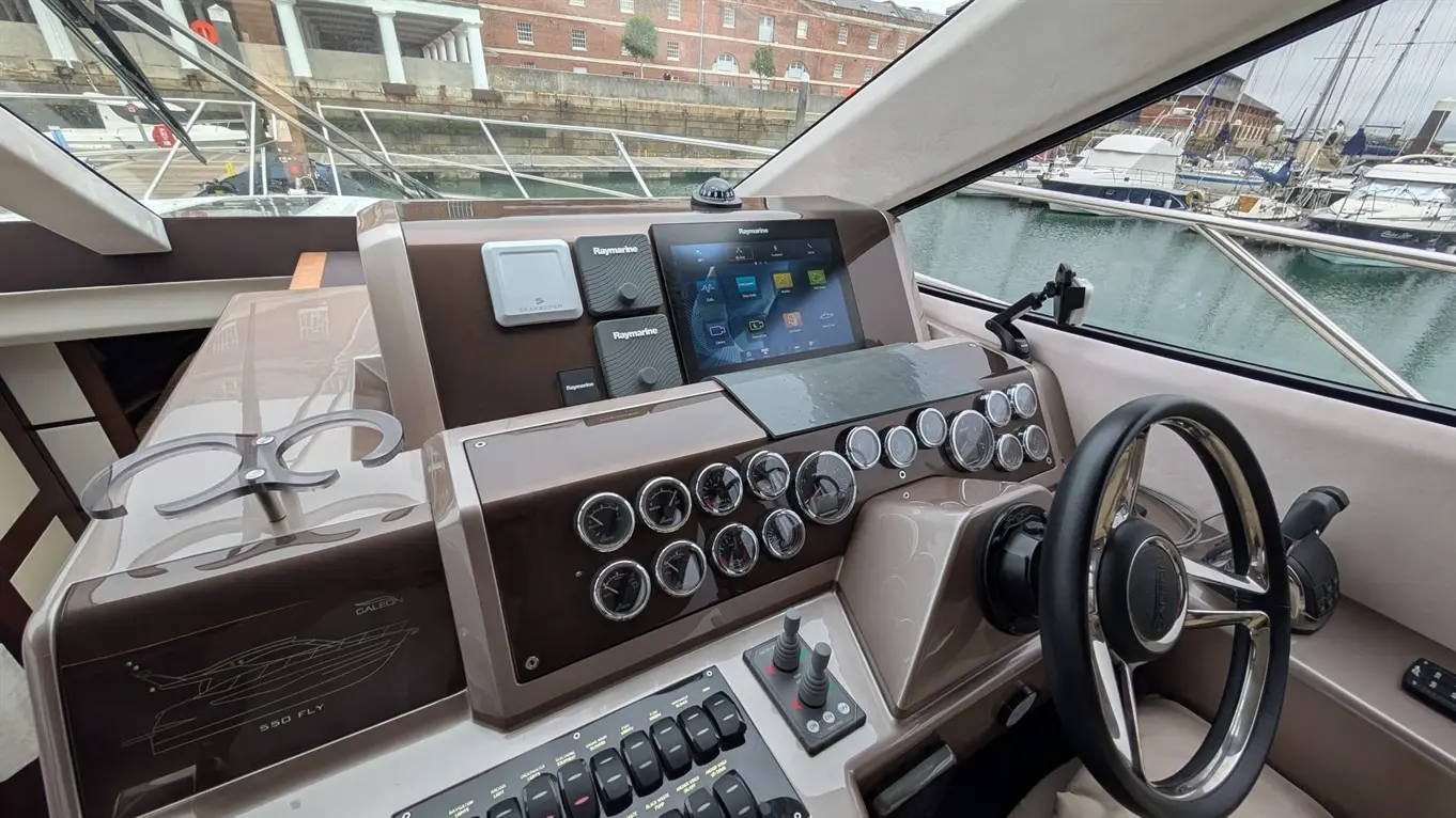 Boat cockpit with a Raymarine touchscreen, gauges, steering wheel, and controls, overlooking a marina with moored yachts.