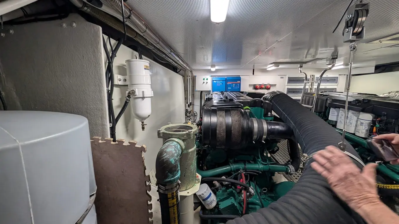 Engine room showing a large green engine, pipes, and control panel; a person's hand is adjusting a black hose.