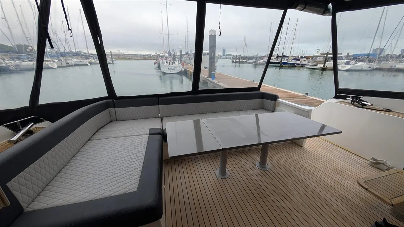 Stylish boat interior with grey seating, a smooth table, and a view of a marina with moored boats and overcast skies.