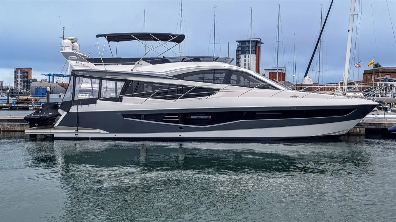 A sleek Galeon 550 yacht docked at a marina, surrounded by other boats, with masts and buildings in the background.