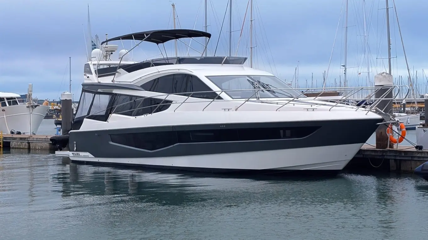 A modern white and grey yacht docked at a marina, reflecting on the calm water, with masts of nearby boats in the background.