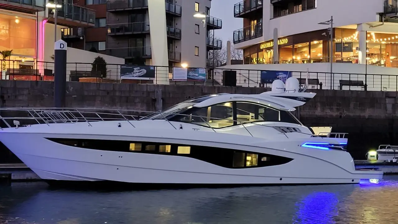 A luxury white motor yacht docked by a marina, illuminated by blue lights, near modern buildings and restaurants.