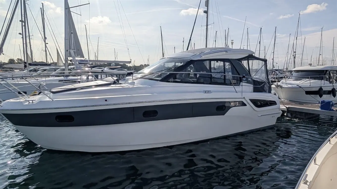 Bavaria S33 HT motor yacht moored in a marina among sailing boats on calm water.