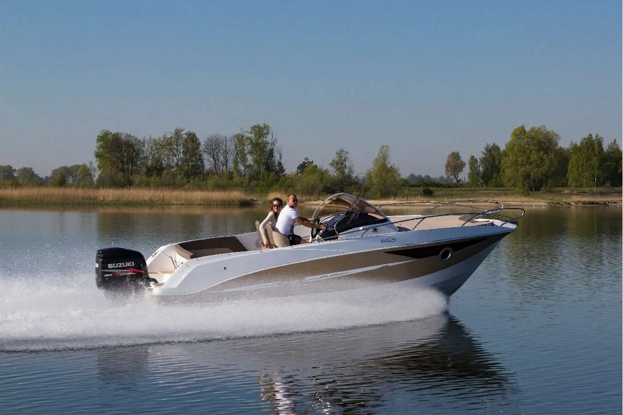 A speedboat with a Suzuki motor cruising on calm water, carrying a man and woman, surrounded by greenery and blue sky.