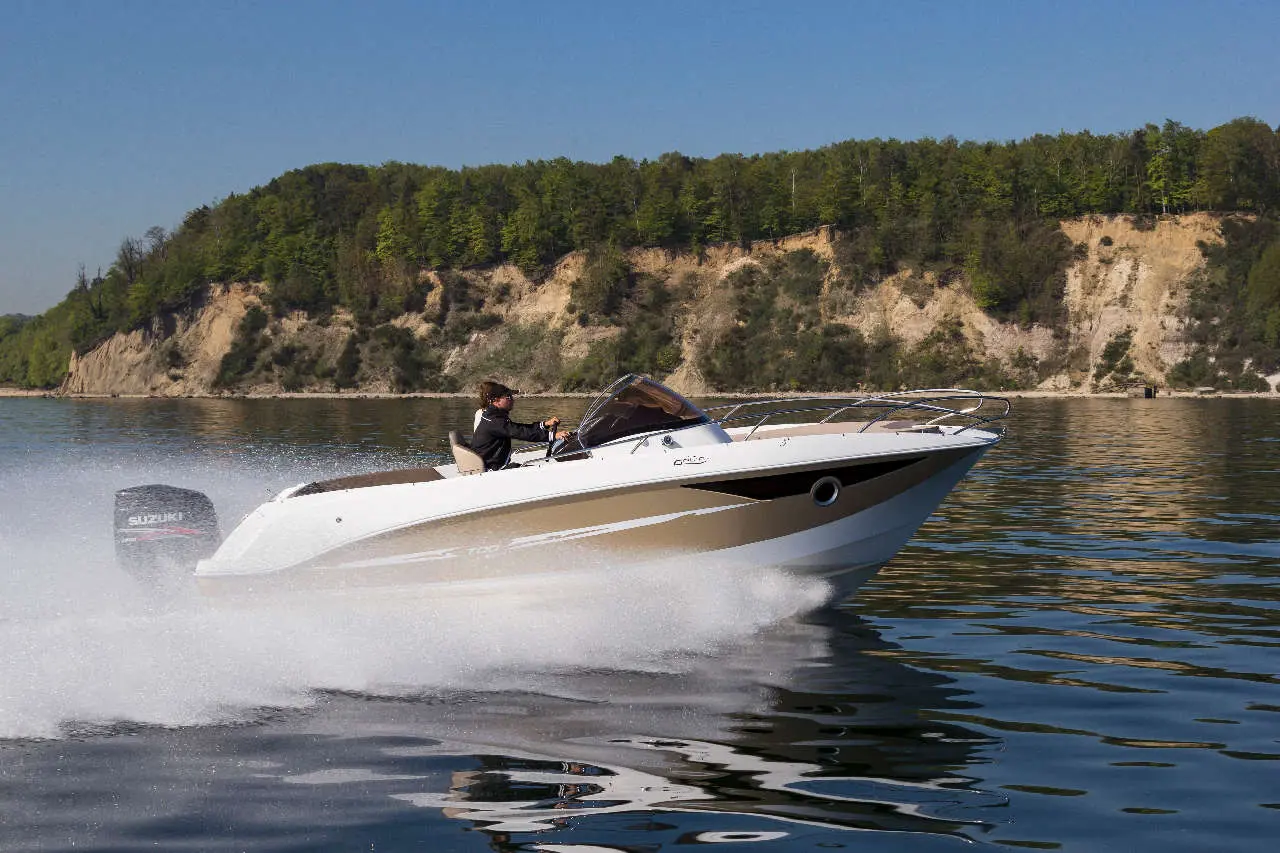A motorboat speeds across calm water, creating a spray, with green cliffs in the background on a sunny day.