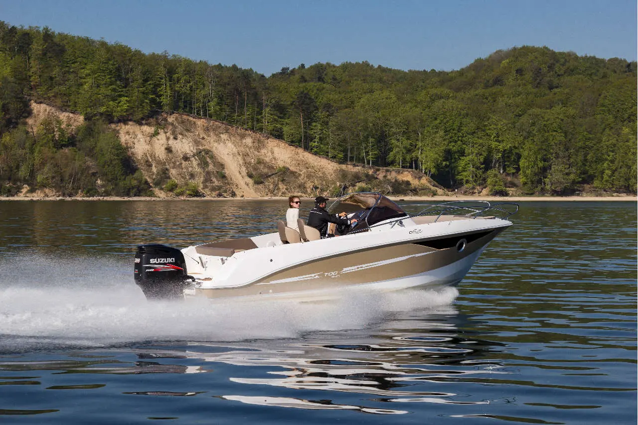 A white and tan speedboat with two people navigates a calm lake surrounded by green trees and a sandy shoreline.