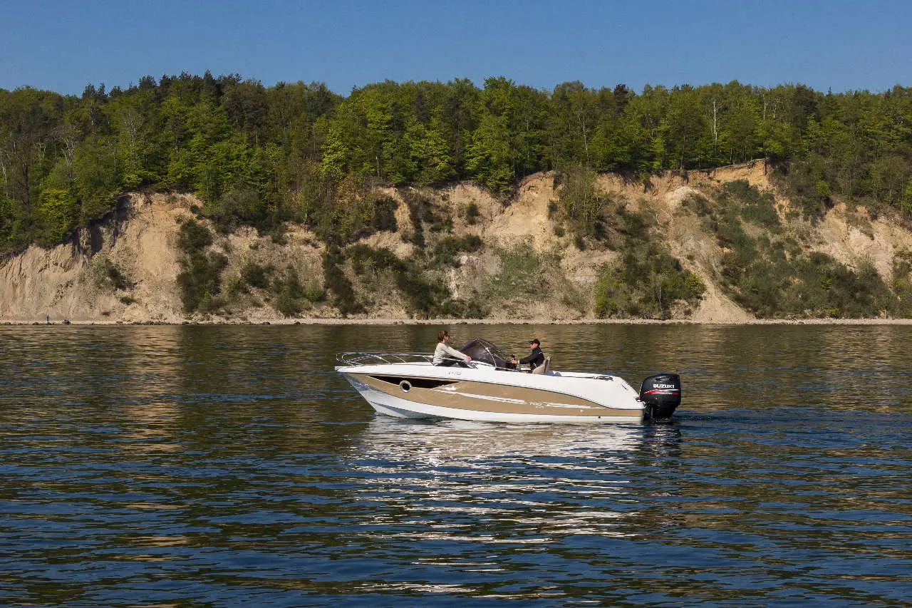 A boat with two people navigates on calm water, surrounded by green trees and a cliff in the background.