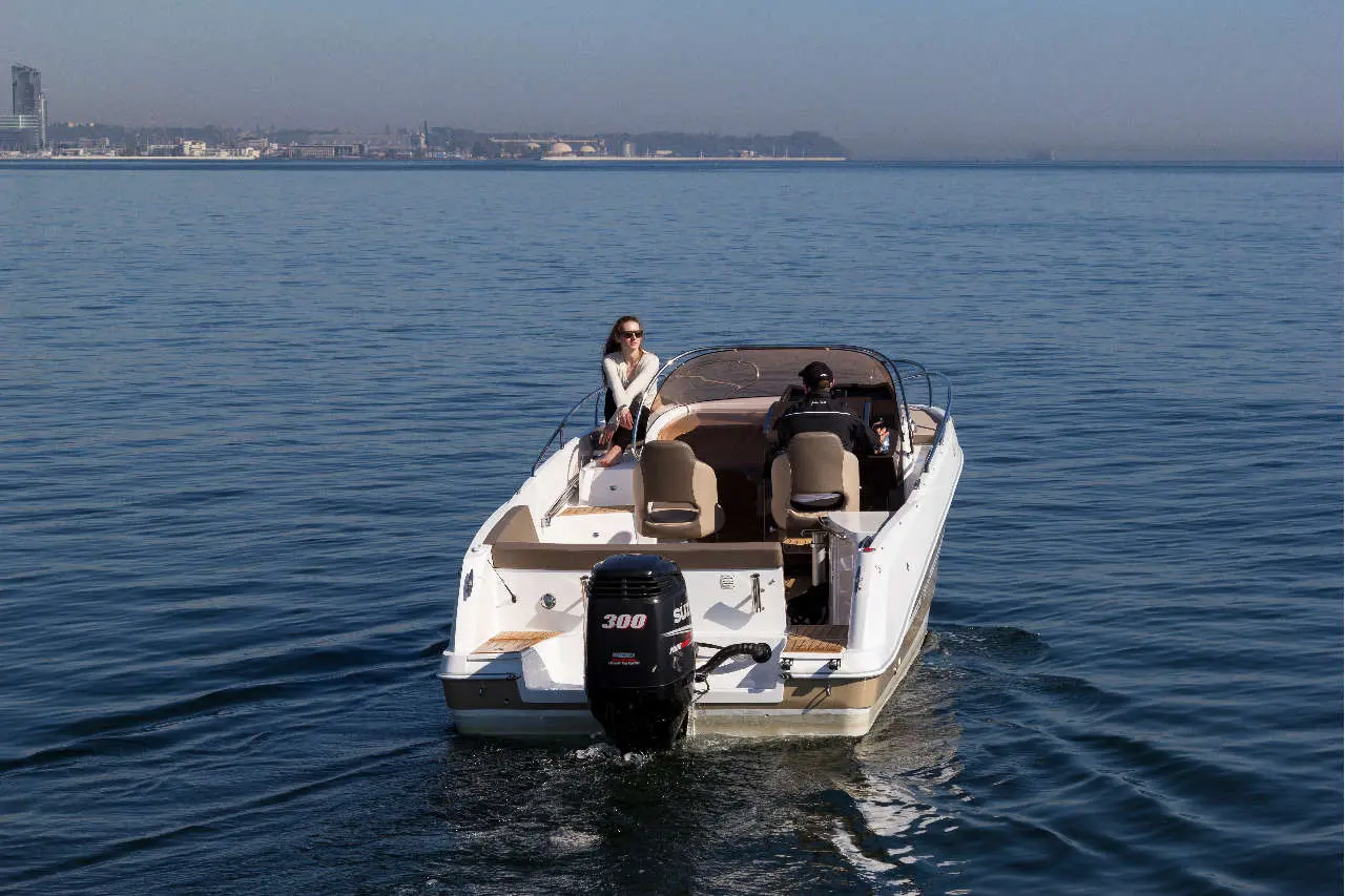 A motorboat with two people on board, cruising on calm water near a city skyline in the background.