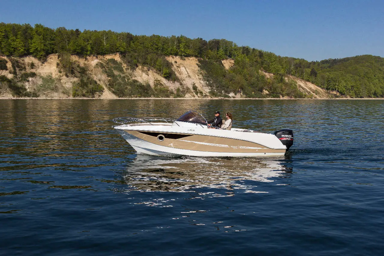 A speedboat glides across calm waters, with two people enjoying a scenic view of green hills and cliffs in the background.