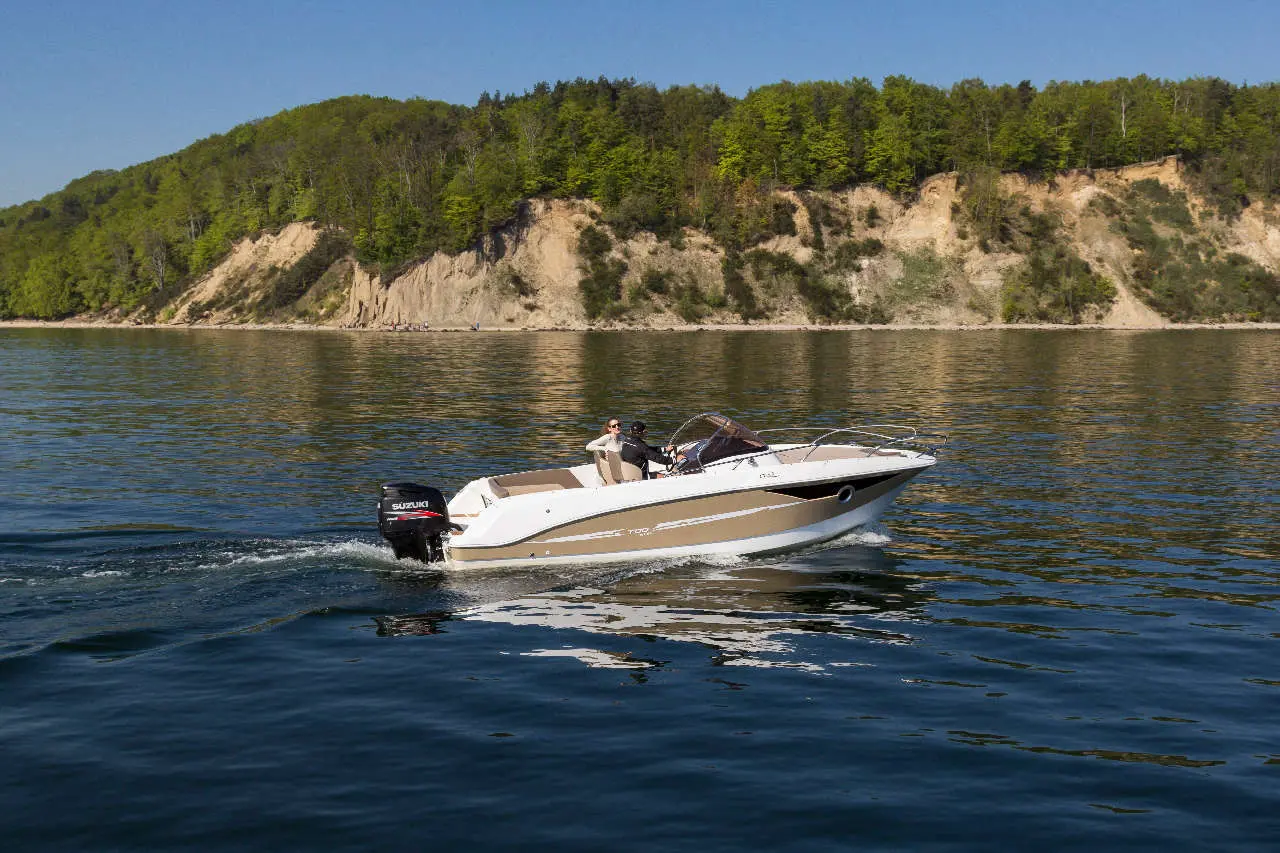 A white and beige boat with two people cruises on calm water near a green wooded shoreline. Suzuki outboard motor visible.