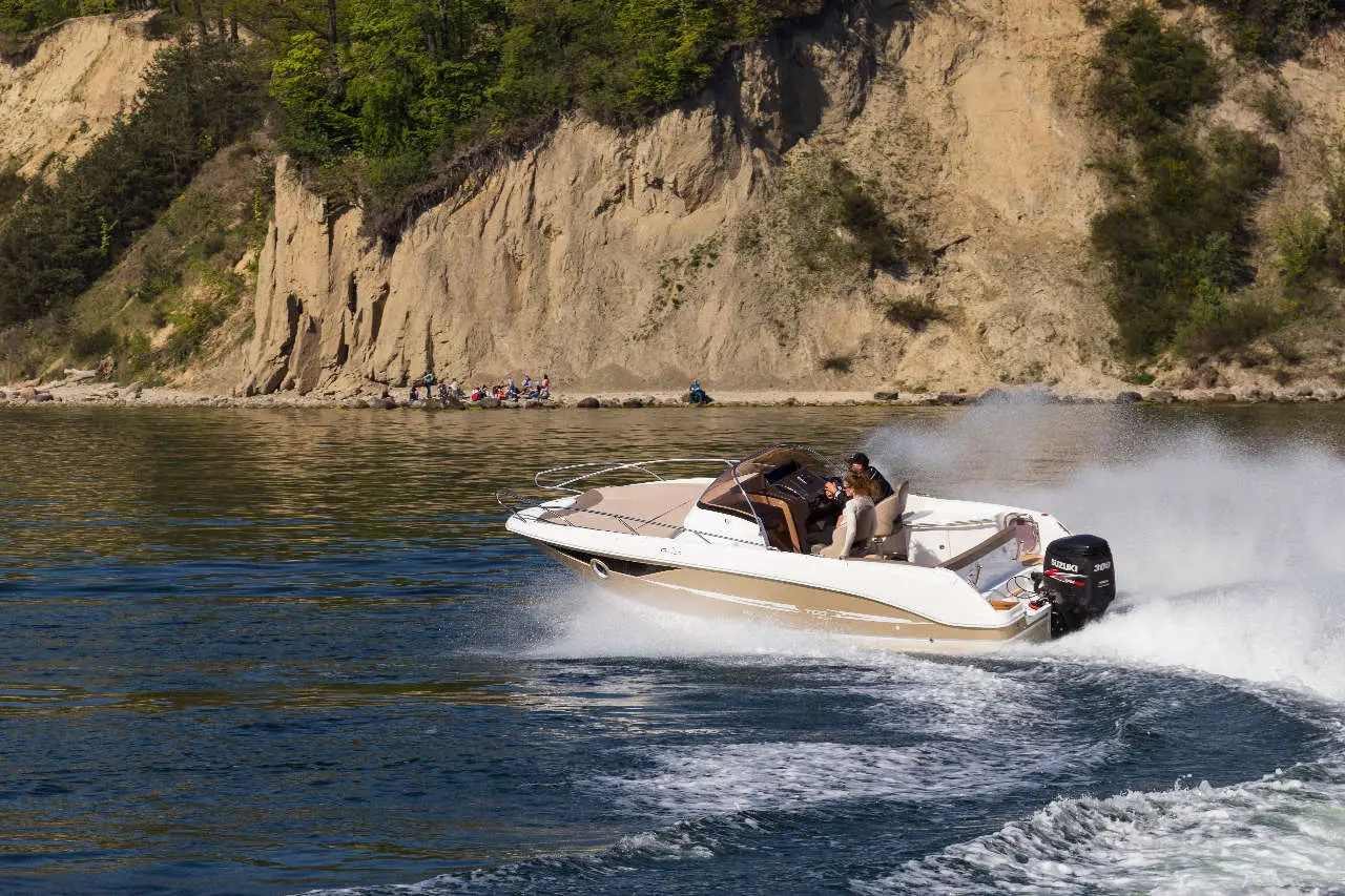 A powerboat with two people speeds across a serene lake, surrounded by rocky cliffs and lively beachgoers in the background.