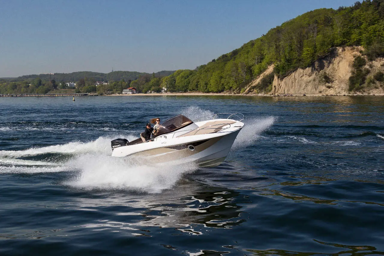 A motorboat speeds through the water, creating splashes, with two people aboard against a scenic riverside background.