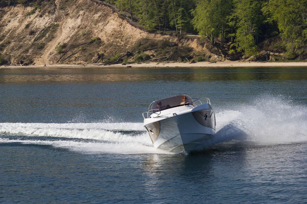 A speedboat turns sharply on calm waters, creating a spray, with a sandy shoreline and greenery in the background.