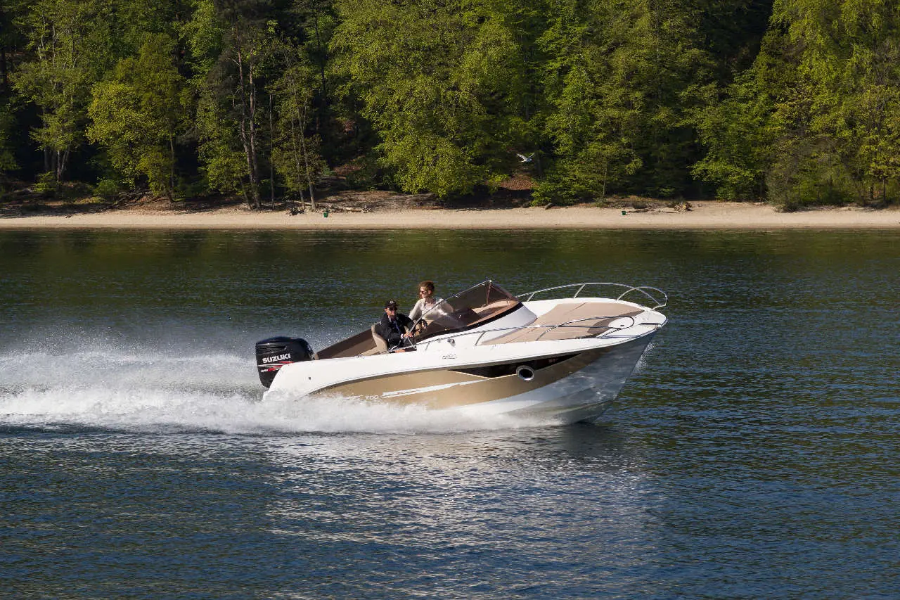 A speedboat with two people glides across calm water, surrounded by green trees and a sandy beach in the background.