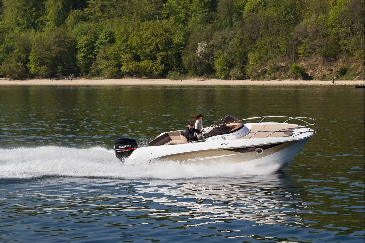 A speedboat with two people cruises on calm water, surrounded by lush greenery and a sandy beach in the background.