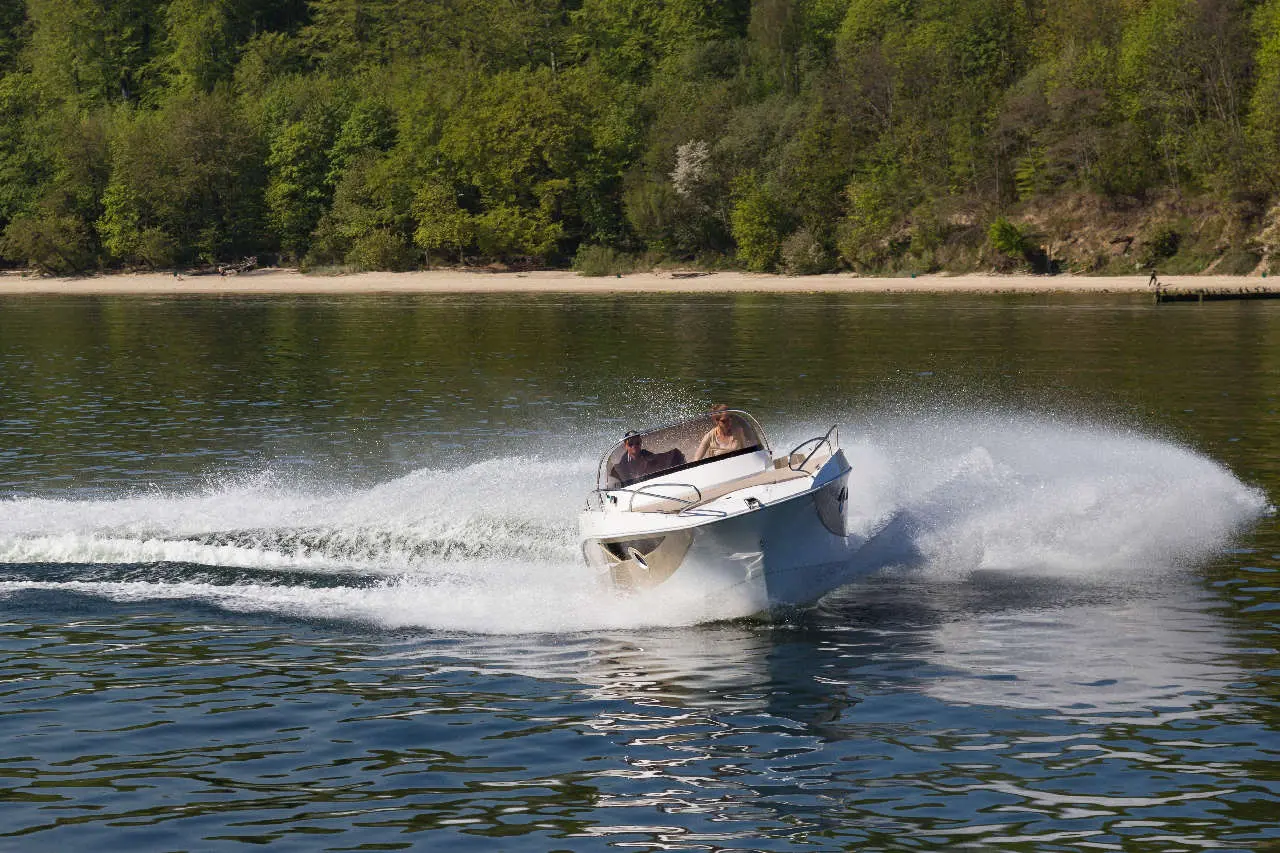 A white speedboat skims across a calm lake, creating spray, with lush greenery and a sandy beach in the background.