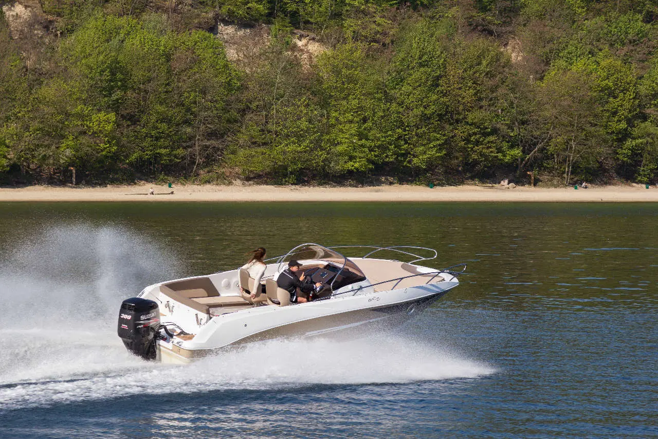 A speedboat with two passengers skims across calm water, leaving a spray behind, with a green landscape in the background.