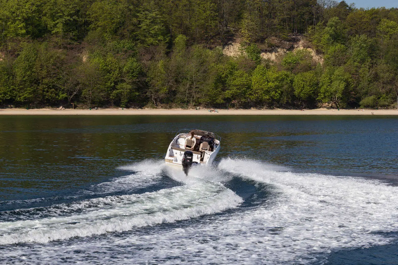 A speedboat creates white wake as it accelerates across a calm lake, surrounded by lush green trees and a sandy shore.