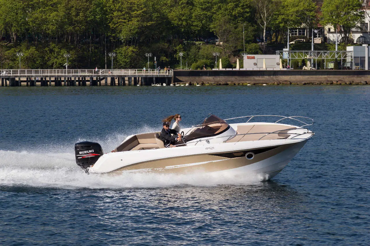 A speedboat with two people speeds across a calm lake, creating waves behind it. A dock and greenery are visible in the background.