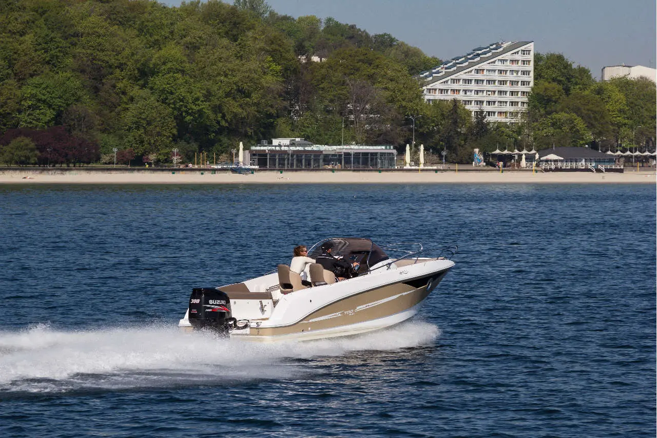 A speedboat with two passengers navigates the water near a beach lined with trees and a modern building in the background.