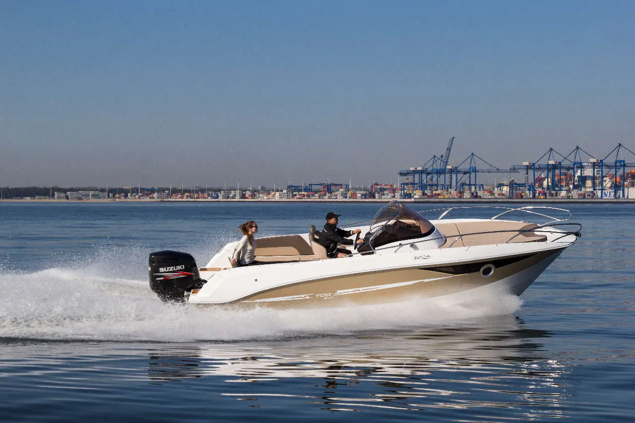 A powerboat, with a driver and passenger, speeds across calm waters near a busy dock and shipping containers.