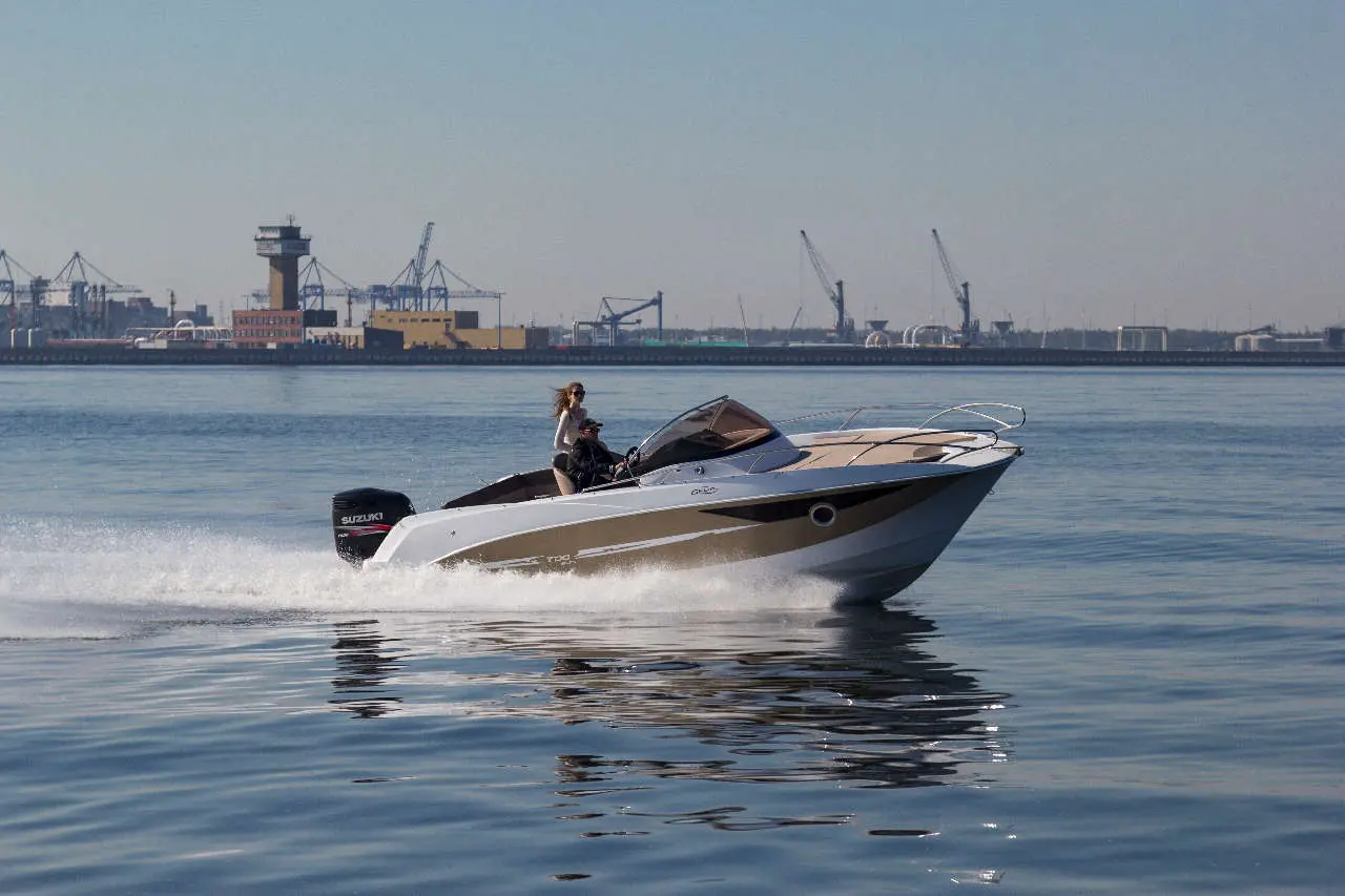 A speedboat with two passengers moves swiftly across calm water, with a port and cranes visible in the background.
