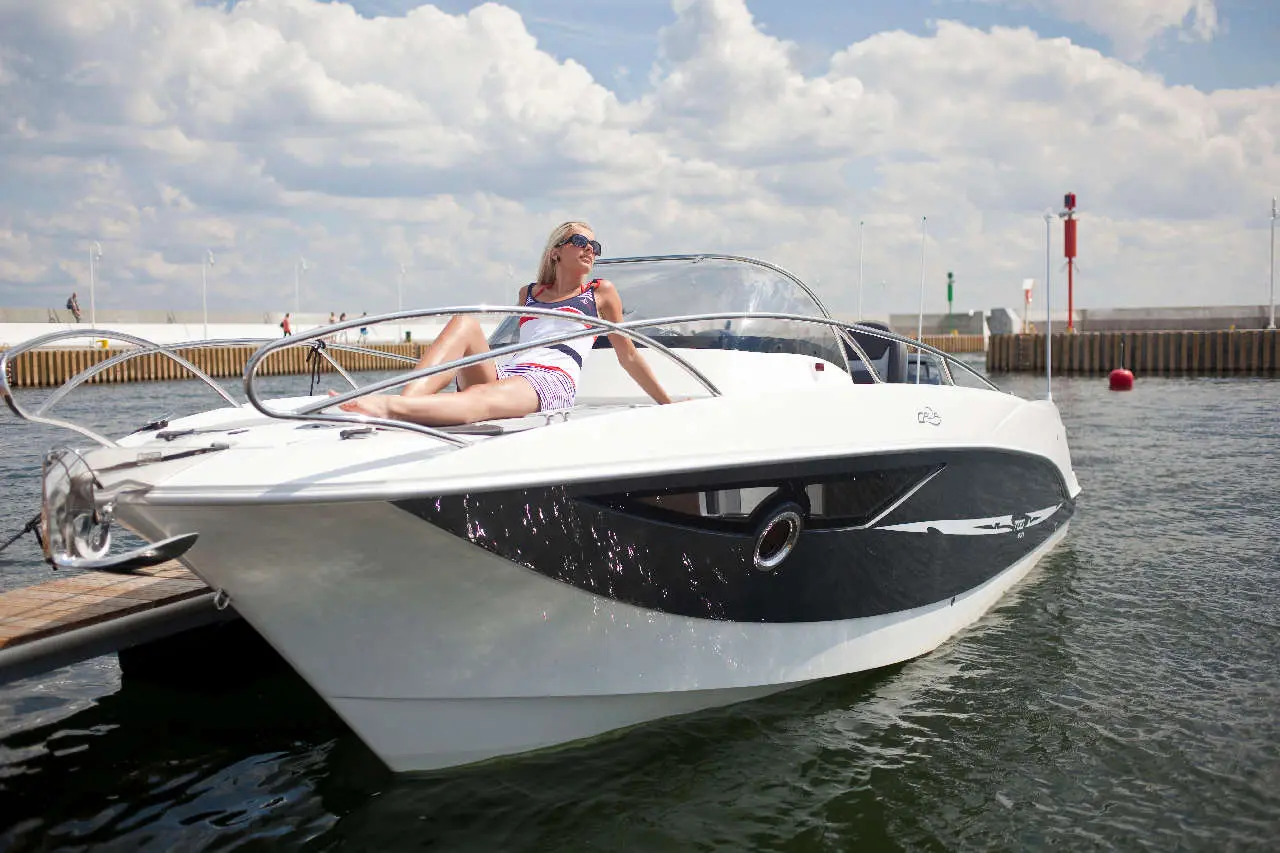 Woman in sunglasses relaxing on a stylish speedboat by the water, with a scenic background of clouds and a marina.
