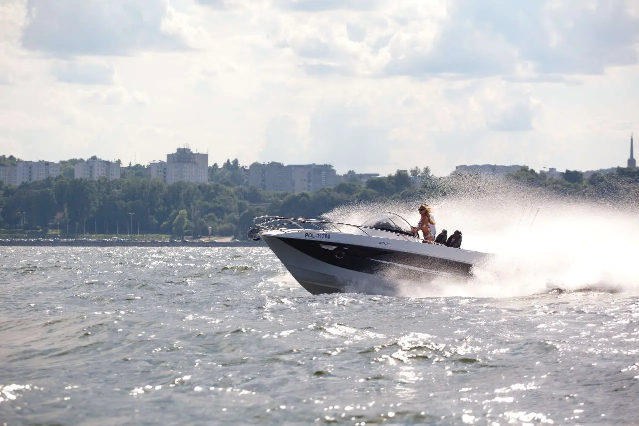 A speedboat with a woman driving skims across a shimmering lake, with a city skyline and trees in the background.
