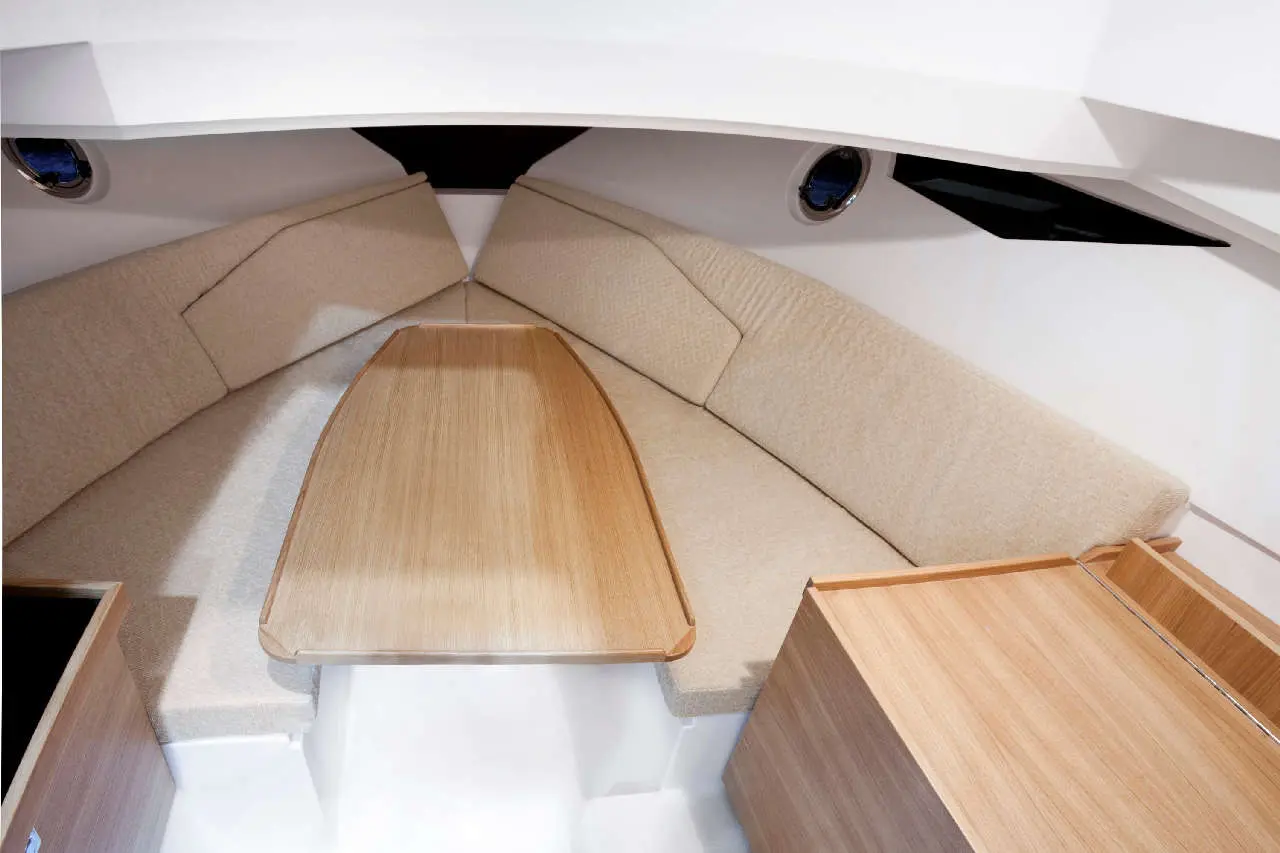 Interior of a boat featuring cream upholstery, a wooden table, and storage, with circular portholes letting in natural light.